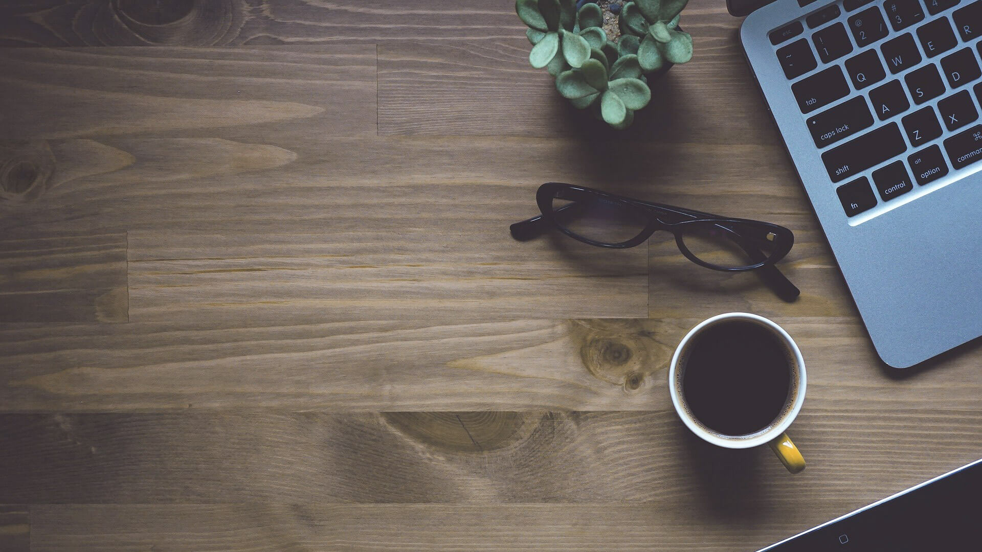 Auf einer Schreibtisch eines Texters befinden sich eine Brille, ein Becher mit Kaffee, ein Laptop und eine Grünpflanze.