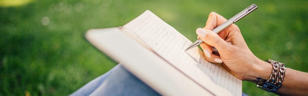 Eine Frau sitzt auf einer Wiese und macht handschriftliche Notizen in ein Notizbuch.