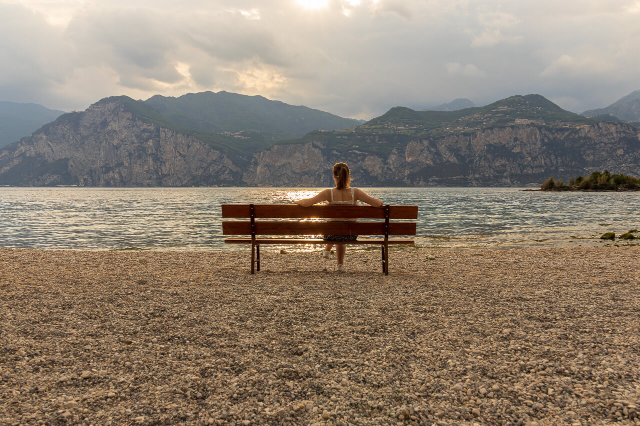 Eine Frau sitzt am Gardasee und blickt bei Sonnenuntergang auf die Berge.